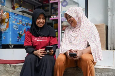 two women with headscarves sit on the curb outside a shop while looking down at their mobile phones