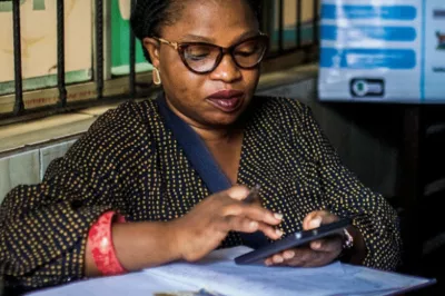 Using her mobile phone, OLUBAM Enterprises’ digital finance agent Ajayi Yemisi completes a mobile money transaction for a customer – Akande Ibrahim – in her shop in the Ladi-Lak area of Bariga in Lagos, Nigeria. 